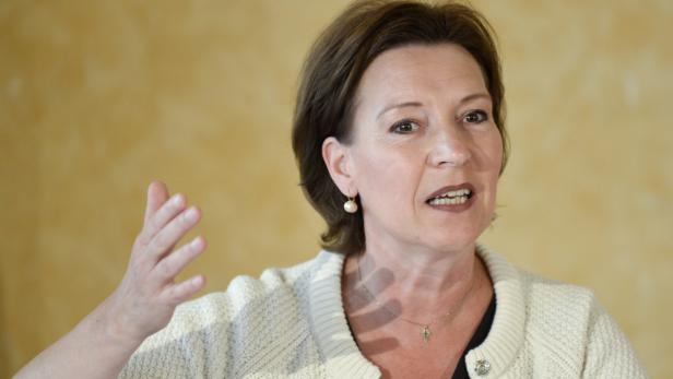 Eine Frau mit kurzem braunem Haar gestikuliert vor einem gelben Hintergrund.