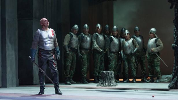 Ein Mann in Rüstung steht vor einer Gruppe von Soldaten auf einer Theaterbühne.