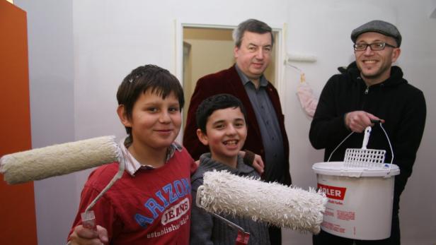 Zwei Jungen, ein Mann mit Farbeimer und ein weiterer Mann mit Farbroller stehen in einem Raum.