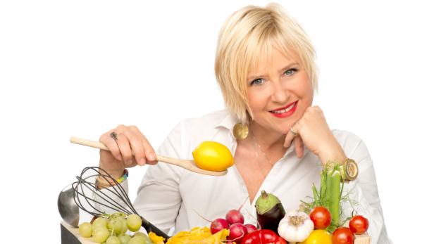 Eine lächelnde Frau mit blonden Haaren präsentiert Gemüse und Obst auf einem Tablett.