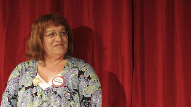 Eine Frau mit Brille und einem Button „Stop Obłudzie“ vor einem roten Vorhang.