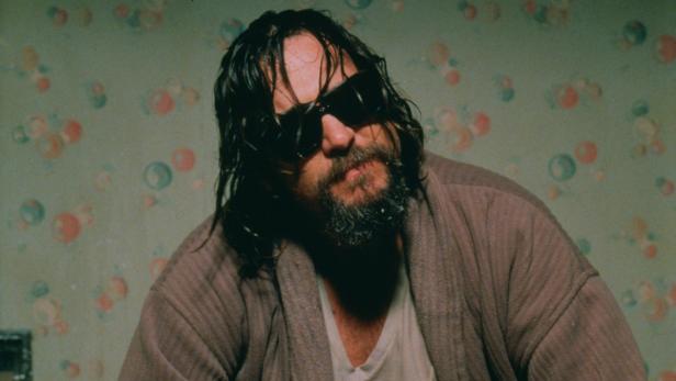 Ein Mann mit Sonnenbrille, Bart und Bademantel vor einer gemusterten Tapete.