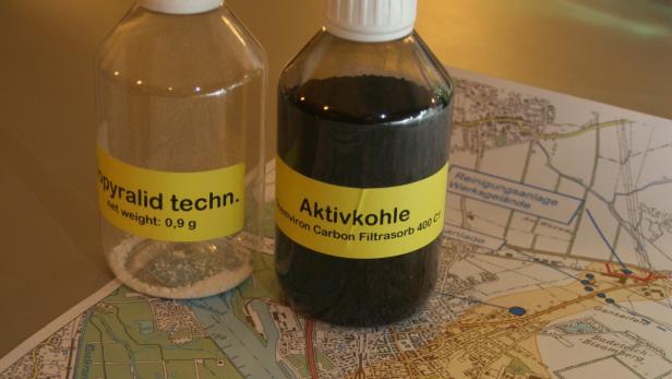 Zwei Flaschen, eine mit Opyralid und eine mit Aktivkohle, stehen auf einer Landkarte.