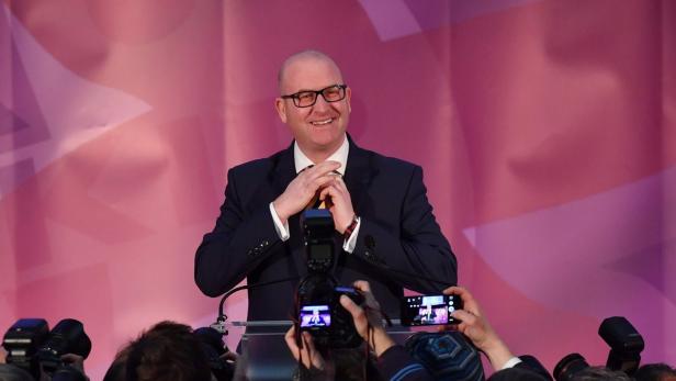 Ein Mann mit Brille steht lächelnd vor einem Publikum und einer pinkfarbenen Wand.