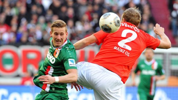Ein Fußballspieler in Rot tritt gegen einen Ball, während ein Gegenspieler in Grün zusieht.