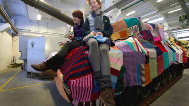 Zwei Frauen sitzen auf einem mit bunten Strickarbeiten bedeckten Panzer.