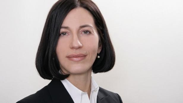 Eine Frau mit dunklen Haaren und einem schwarzen Blazer vor einem hellen Hintergrund.