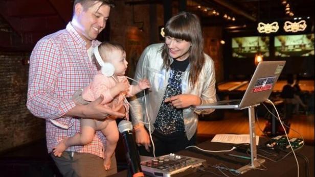 Ein Mann hält ein Baby mit Kopfhörern, während eine Frau auf einen Laptop und ein Mischpult schaut.