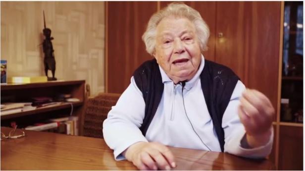 Die 89-jährige Gertrude die hinter einem braúnen Tisch auf einem Sessel sitzt