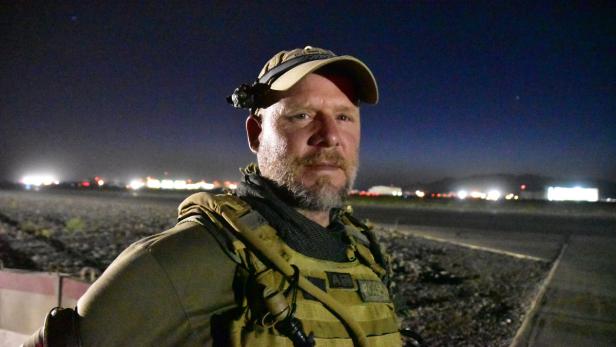 Ein Mann mit Bart und taktischer Ausrüstung steht nachts auf einem Flugfeld.