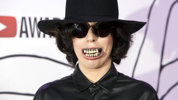 Lady Gaga mit Hut und auffälligem Zahnschmuck bei einer Preisverleihung.
