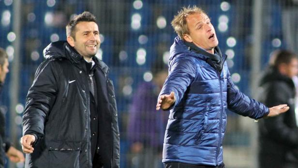 Zwei Fußballtrainer stehen am Spielfeldrand, der eine lächelt, der andere gestikuliert aufgeregt.
