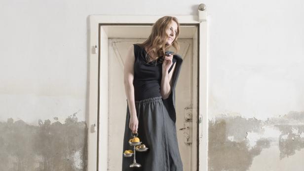 Eine Frau in einem schwarzen Top und einem grauen Rock hält eine Etagere mit Zitrusfrüchten vor einer Tür.