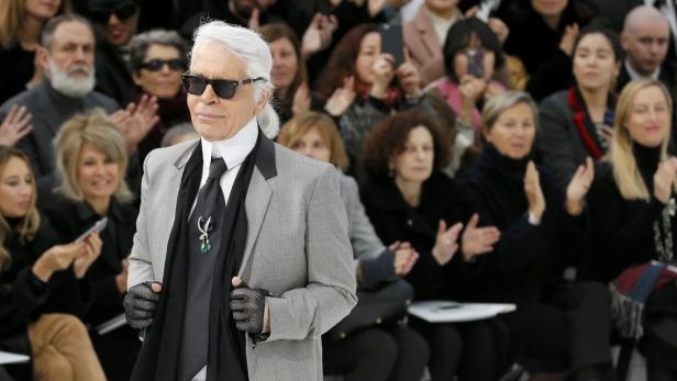 Karl Lagerfeld bei einer Modenschau, mit Sonnenbrille und Handschuhen, vor einem applaudierenden Publikum.