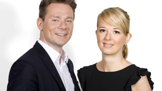 Ein Mann im Anzug und eine Frau im schwarzen Kleid lächeln in die Kamera.