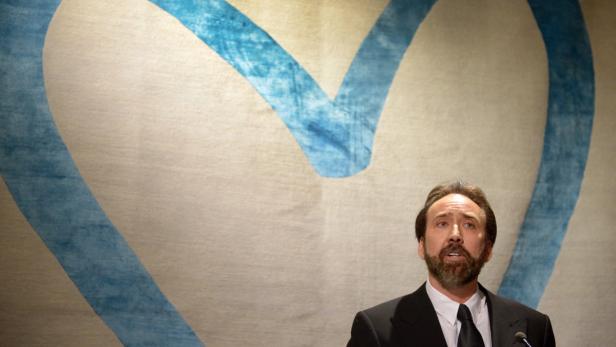 Nicolas Cage steht vor einem Wandteppich mit einem blauen Herz.