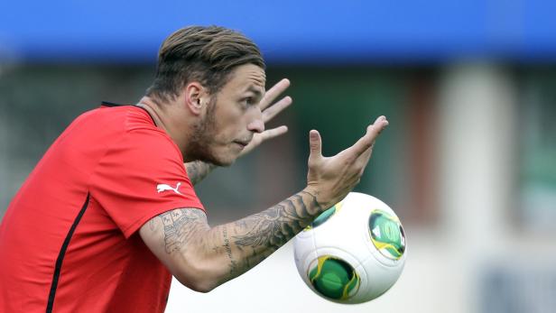 Ein tätowierter Mann in rotem Trikot fängt einen Fußball.
