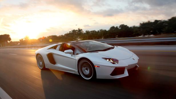 Ein weißer Lamborghini fährt auf einer Straße in Richtung Sonnenuntergang.