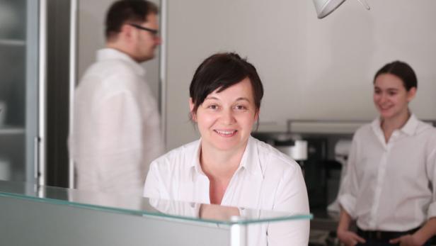 Eine lächelnde Frau steht hinter einem Tresen, im Hintergrund zwei weitere Personen.