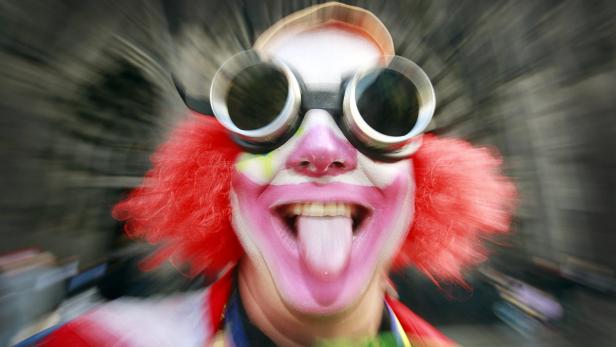Ein Clown mit roter Perücke und übergroßer Brille streckt die Zunge heraus.