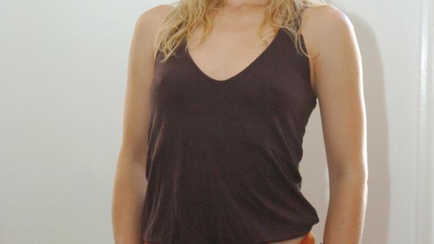 Eine Frau mit blonden Haaren, braunem Top und orangefarbener Hose posiert vor einer weißen Wand.