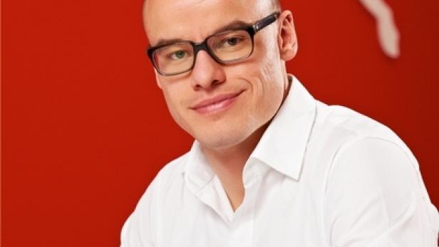 Ein Mann mit Brille und weißem Hemd posiert vor einem roten Hintergrund mit dem Puma-Logo.