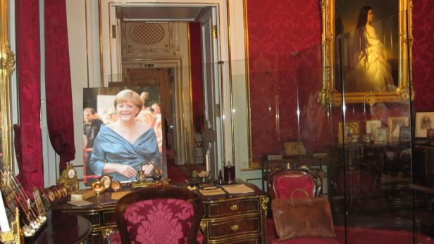 In einem opulenten Raum steht ein Schreibtisch mit einem Porträt von Angela Merkel.