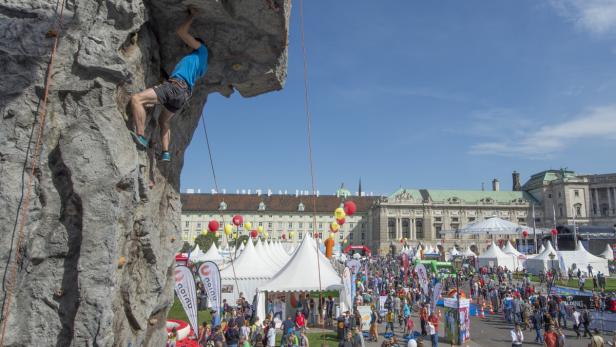 Ein Mann klettert an einer künstlichen Felswand vor dem Hintergrund eines belebten Stadtfestes.