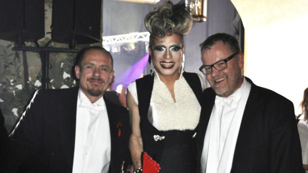 Drei Personen, darunter eine Dragqueen, posieren lächelnd für ein Foto.