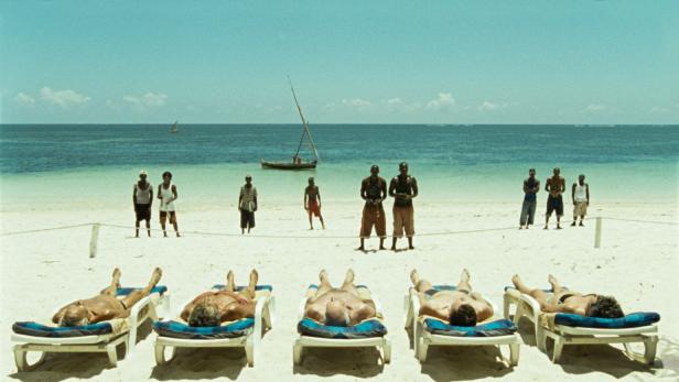 Menschen entspannen sich am Strand, während andere im Hintergrund stehen und ein Segelboot auf dem Wasser fährt.