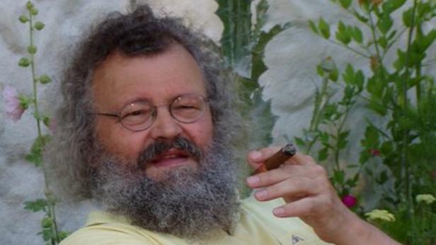 Ein Mann mit Bart und Brille raucht eine Zigarre vor einem Hintergrund aus Pflanzen.