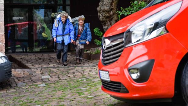 Reinhold Messner und ein Begleiter verlassen ein Gebäude mit Rucksäcken, im Vordergrund ein rotes Auto.