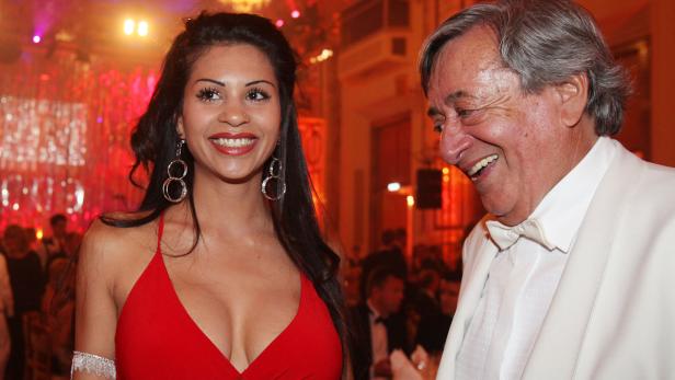 Eine lächelnde Frau in einem roten Kleid und ein Mann im weißen Anzug auf einer Veranstaltung.