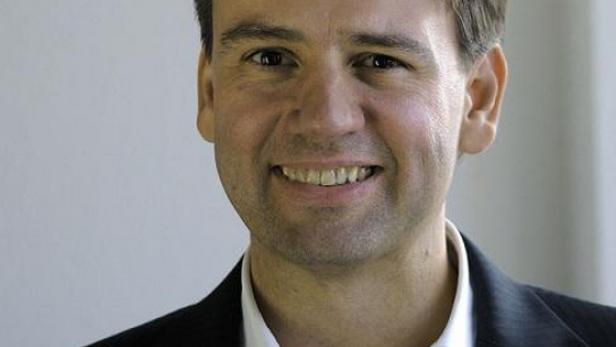Ein lächelnder Mann in einem dunklen Anzug und einem weißen Hemd.