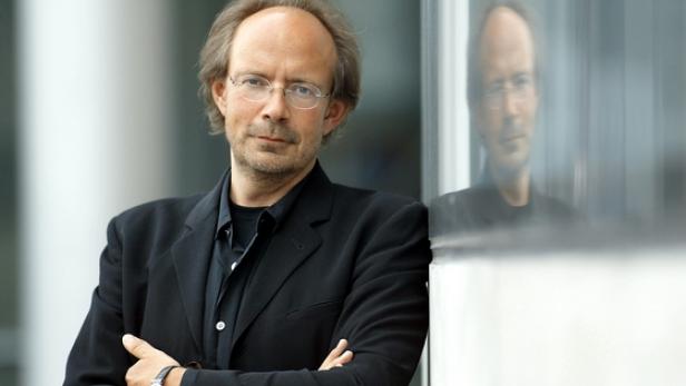 Ein Mann mit Brille und dunklem Anzug steht vor einer Glasfassade.