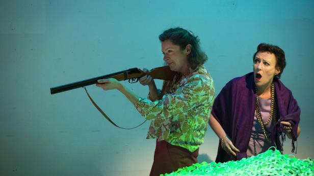 Eine Frau zielt mit einem Gewehr, während eine andere Frau erschrocken daneben steht.