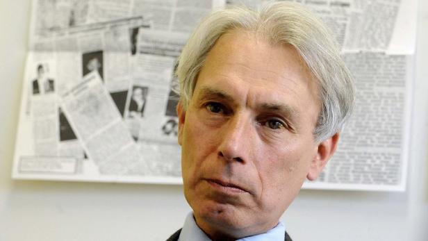 Porträt eines Mannes mit grauem Haar vor einer Wand mit Zeitungsartikeln.