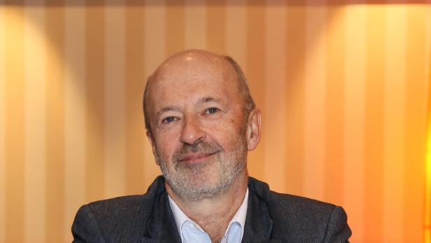 Porträt eines Mannes mit grauem Haar und Bart vor einem gestreiften Hintergrund.