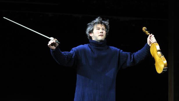 Ein Mann im blauen Pullover hält eine Geige und einen Geigenbogen in den Händen.