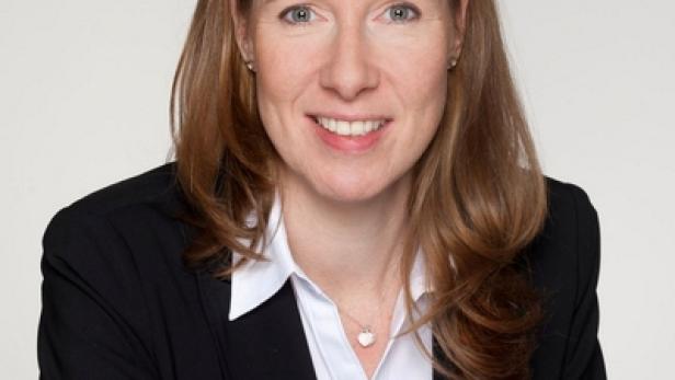 Eine Frau mit braunen Haaren trägt ein schwarzes Jackett und lächelt.