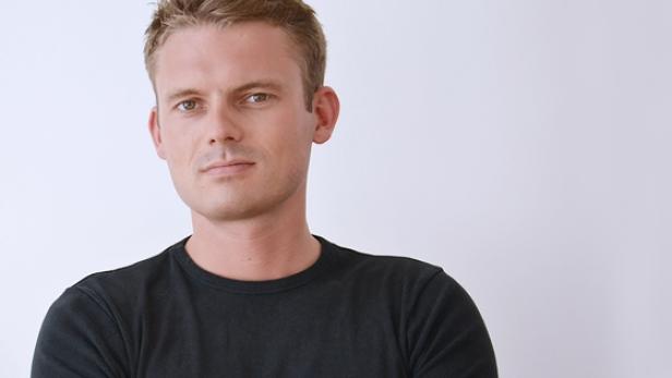 Ein Mann mit blonden Haaren und einem schwarzen T-Shirt vor einem weißen Hintergrund.