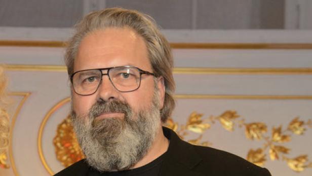 Ein Mann mit Bart und Brille vor einem goldenen Hintergrund.