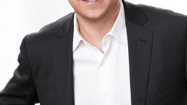 Ein Mann mit braunen Haaren und einem schwarzen Anzug lächelt in die Kamera.