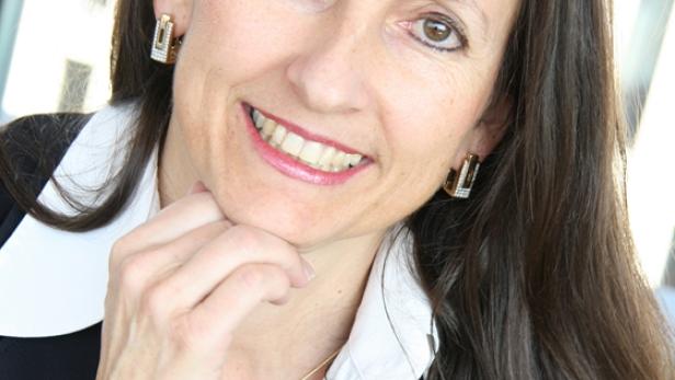 Eine lächelnde Frau in einem Blazer mit weißem Hemd und Schmuck berührt ihr Kinn.