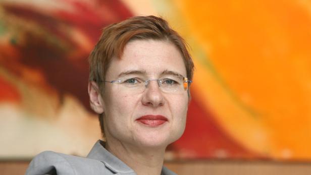 Eine Frau mit Brille und kurzem, rötlichem Haar vor einem orangefarbenen Hintergrund.