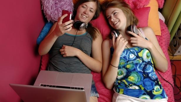 Zwei junge Frauen liegen mit Kopfhörern, Laptop und Smartphone auf einem Bett.