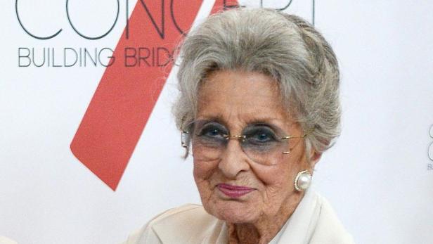 Eine ältere Frau mit Brille und Perlenohrringen lächelt in die Kamera.