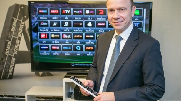 Ein Mann im Anzug hält eine Fernbedienung vor einem Fernseher mit verschiedenen TV-Logos.