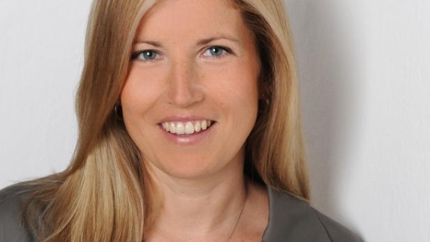 Eine lächelnde Frau mit blonden Haaren und einem grauen Blazer vor einer weißen Wand.