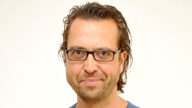 Porträt eines Mannes mit Brille und braunen, gewellten Haaren vor einem hellen Hintergrund.